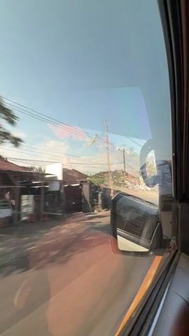 The video appears to be taken from inside a vehicle, looking out the window. The first frame shows a road with some buildings and trees on the side, under a clear blue sky. The second frame shows a large red truck passing by, its details blurred due to motion. The third frame returns to a view of the road, with houses and vegetation lining the side, again under a clear sky. The camera is panning or the vehicle is moving, creating a sense of travel. There are power lines visible in the background throughout the video.