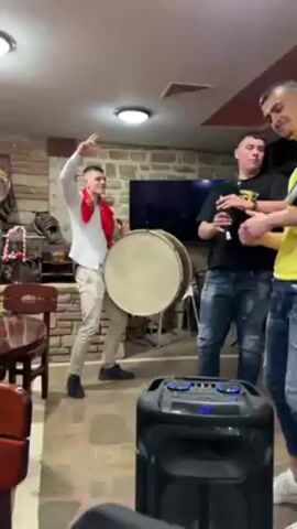 In a brightly lit indoor setting with stone-accented walls and tiled floors, three young men are the focus of the video. The first man, wearing a white shirt, gray pants, and a red scarf, is playing a large drum while dancing energetically. The second man, in a black shirt and jeans, plays a bagpipe-like instrument. The third man, wearing a yellow t-shirt and jeans, holds a smaller drum and gestures with his hands. Between them is a large black speaker with illuminated buttons. In the background, there's a dark wooden table and chairs, a partially visible bar area with decorative items, and a television. The scene suggests a lively, possibly traditional, musical performance.
