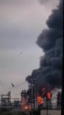 This video captures a significant industrial incident at a refinery in Villeneuve-le-Roi, France. The primary subject is the refinery itself, a complex of industrial structures including tall stacks, pipes, and elevated walkways. The scene is dominated by a large, intense fire erupting from one of the refinery units, sending thick, dark smoke billowing into the sky. Flames are clearly visible, licking up from the industrial equipment.

The activity taking place is a major fire at the refinery, suggesting an emergency situation. Water is being sprayed from a tall structure, possibly an aerial ladder truck or an integrated firefighting system within the refinery, indicating an attempt to combat the blaze. The weather appears overcast, with a grey sky, which contrasts with the bright orange and red of the flames. The time of day is not explicitly discernible, but the overcast conditions might suggest daylight hours.

Notable details include the scale of the fire and smoke plume, which are substantial and visually impactful. The presence of what appears to be a bird or drone in the upper left portion of the frame is a minor detail. The background includes a residential building, highlighting the proximity of the industrial facility to civilian areas. There is no visible text within the video frames. The overall emotion conveyed by the imagery is one of urgency and potential danger due to the industrial accident.