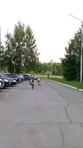 A boy rides a bicycle on a paved surface with cars parked on the left and trees and grass on the right. He performs a wheelie, balancing on the back wheel. Another boy on a smaller bicycle follows behind him. The boy doing the wheelie smiles at the camera as he brings the bicycle down.