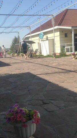The video captures an outdoor scene, likely a residential backyard or a resort area in Dobun, Kazakhstan. The setting features a paved stone patio, a building with a reddish-brown roof and beige siding, and lush green trees. The time of day appears to be sunny and warm, with clear blue skies and noticeable shadows, suggesting midday.

In the foreground, a person is seen on a swing, while another person stands nearby. A decorative circular swing, adorned with artificial greenery and red flowers, hangs from a white metal structure, suggesting a festive or recreational atmosphere. A string of colorful lights hangs overhead, adding to the ambiance.

The building in the background has large windows and a porch area, hinting at a place for relaxation or living. There are several potted plants and garden decorations scattered around the yard.

A blue satellite dish is visible on the side of the building. Two signs are attached to the white metal structure, but the text is too blurry to read clearly. One sign has "#family_resort" written on it.

The overall mood of the video seems peaceful and relaxed, with people enjoying the outdoors. The clean and well-maintained appearance of the area suggests a pleasant environment.