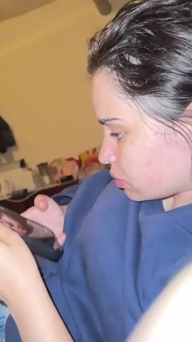 A young woman is holding a phone in her left hand and looking at it. Her right hand is also visible, holding the phone. She has dark hair and is wearing a dark blue sweatshirt. Her face is turned to the side at first, then she turns to look at the camera and smiles. The background is out of focus and appears to be an indoor setting.