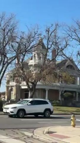The video shows an exterior shot of a large, historic-looking house with a turret. Trees with bare branches partially obscure the view of the house. The house appears to be located in a residential area, as there's a street and a yellow fire hydrant visible in the foreground. A white SUV is partially visible on the left side of the frame. The sky is blue, suggesting a clear day. The house has decorative details and a porch. There are also some evergreen decorations on the porch.
