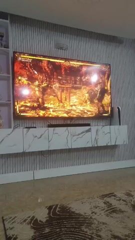 A person is playing the video game Mortal Kombat on a large, wall-mounted flat-screen television. The characters in the game are fighting in a fiery environment with trees and traditional Japanese architecture in the background. The television is mounted above a white console with a faux marble finish, positioned against a textured gray wall with vertical ridges. There is a sound bar located on the console beneath the television. The floor is shiny and patterned with dark, organic shapes.