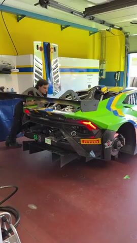 The video shows a man in a garage working on a green, blue, and yellow race car. The car, with the number 63 and "PRO" displayed, is on jack stands with its rear wheels off, revealing the brake rotors. The man is focused on the rear of the vehicle, near the large rear wing. A mechanic's tool is visible nearby, and the walls of the garage are painted in a yellow and blue scheme, adding to the bright and functional atmosphere of the space.