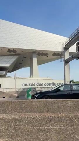The video opens with a close-up of the base of a large, weathered concrete or stone wall, likely part of an overpass or structure. Several small, dark, bulbous objects are lined up on the ground at the foot of the wall. The camera then pans up and to the right, revealing a highway with traffic.

In the background, a striking modern building with a geometric, angular design and a grey/silver facade is prominent; this is the Musée des Confluences. A road sign indicating directions to "GERLAND", "BERGES DU RHONE", and "LA CONFLUENCE" is visible. Cars and a large white semi-trailer truck are seen driving on the highway. On a concrete barrier next to the road, the words "musée des confluences" are clearly visible, along with some graffiti. The sky is clear and blue, indicating a sunny daytime setting. The camera continues to pan, following the vehicles on the highway.