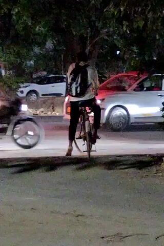The video shows a person on a bicycle at night. The person is wearing a backpack and dark pants. They are riding on a road with traffic. Cars and a motorcycle are visible in the background, some with their headlights on, creating motion blur. Trees are present along the side of the road. The lighting is dim, with streetlights and vehicle lights illuminating the scene. The cyclist appears to be moving forward.