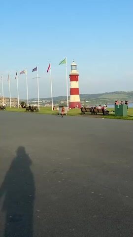 The video shows a park setting with a red and white lighthouse in the background. There are multiple flagpoles with various flags. People are sitting on benches and walking around. A child in a red shirt runs towards the benches. The park overlooks the sea. The sky is clear and blue. The camera pans to the right, showing more of the park and people enjoying the scenery. There's a lamp post and a waste bin near the benches. Some people are sitting on the grass, and a stroller and a scooter are parked next to a bench.
