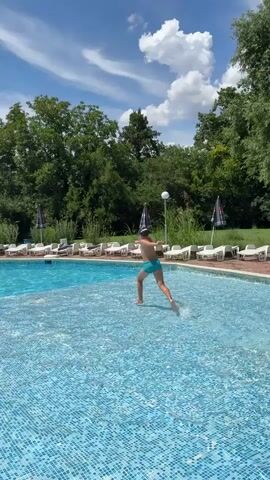 A person in blue swim trunks jumps into a swimming pool. The person lands in the water, creating a splash, and then emerges from the water, looking at the camera. The pool is surrounded by lounge chairs and umbrellas. In the background, there are trees and a blue sky with clouds.