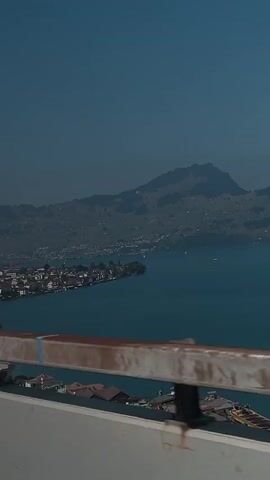 The video captures a view from a moving vehicle, likely a car or bus, looking out over a vast blue lake towards a distant mountainous landscape. In the foreground, a weathered metal guardrail with reddish-brown markings runs horizontally across the lower portion of the frame. The lake is calm, with a few small boats visible on its surface. Along the left shore of the lake, a town or village is nestled, characterized by its red-tiled roofs and buildings that hug the shoreline. The landscape beyond the town rises into rolling hills and then culminates in larger mountains that dominate the horizon. The sky is a clear, deep blue. The overall impression is one of a scenic drive through a picturesque, possibly European, location.