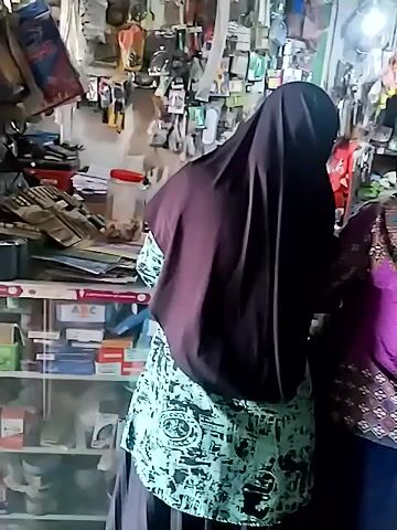The video shows two women in a store. Both women are wearing hijabs, covering their heads and faces, and long dresses. The woman on the right is wearing a pink and brown patterned dress. The woman on the left is wearing a black dress. The store appears to be cluttered with shelves stocked with various items. The camera angle is high and looking down at the women. The lighting is dim.