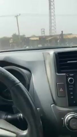 The video, shot from the driver's seat of a car, shows a drive through a rural or developing area.  The camera is static, capturing the view through the windshield. Initially, the car is stationary, possibly at an intersection or waiting to merge. A man stands by the roadside, near some simple structures, likely shops or homes. As the car starts moving, it drives down a paved road. The road is bordered by blue and yellow bollards, suggesting a delineation or barrier. Other vehicles are visible in the distance, traveling in the same direction.  The overall environment appears dusty or hazy, with tall telecommunication towers visible in the background.