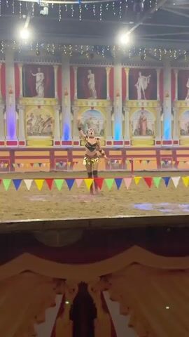 A woman in a gladiator costume is performing in a dirt arena. The costume consists of a bra top with gold accents, a short skirt with gold leaf-like embellishments, gold armbands, and knee-high boots. She is also wearing a headdress with gold accents. The arena is decorated with a backdrop of Roman-style architecture, including columns and painted murals. Colorful triangular flags are strung across the front of the arena. The woman is captured in various poses as she dances or performs, with spotlights of pink and green light illuminating the area. She appears to be smiling and engaged in her performance.