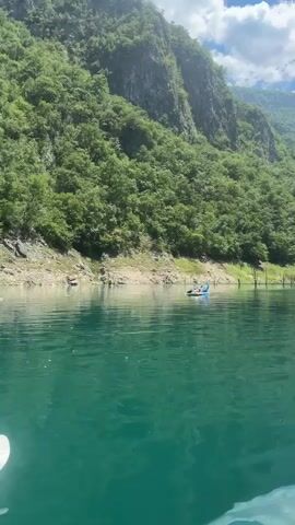 A person is paddleboarding on a tranquil, turquoise lake. The paddleboarder, wearing a blue shirt and white shorts, is lying down on the board, holding their paddle up in the air. The paddleboard is blue and white. The lake is calm and reflects the surrounding lush green mountains. The mountains are covered in dense forest, with rocky cliffs visible in places. The sky is partly cloudy, with white clouds against a bright blue backdrop. The scene appears to be set in a remote, natural environment. The lighting suggests it is daytime, likely late morning or early afternoon. The overall mood is serene and peaceful, conveying a sense of relaxation and enjoyment of the outdoors. There is no visible text in the media file.