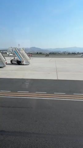The video shows an airport tarmac on a clear day. In the foreground, there is a paved surface with yellow and white markings. In the middle ground, there are several airplanes parked on the tarmac. One airplane, a white turboprop with twin engines, is visible on the left side of the frame, with a set of mobile stairs attached to its fuselage. Further in the distance, other aircraft are parked, and some appear to be in motion. The background consists of a flat, paved expanse of the airport, with buildings and hills visible on the horizon under a clear blue sky. No people are clearly discernible in the frame. The overall scene depicts a typical airport environment.