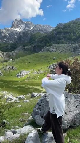 A woman stands on a large rock in a mountainous, grassy landscape with her back to the viewer. She is wearing a white hoodie and brown pants. She raises her arms in the air and yells, "Woo!" In the background, there are snow-capped mountains, a green valley, and a blue sky with scattered clouds. The scene is bright and sunny.