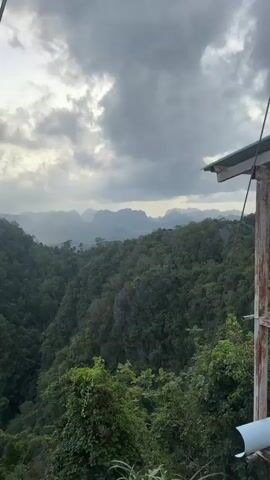 A panoramic video captures a lush green forest landscape with dramatic mountain formations in the distance under a cloudy sky. The foreground is dominated by dense foliage, with glimpses of jagged rocks and a structure partially visible in the upper right corner. The view suggests a high vantage point, overlooking a vast expanse of tropical wilderness. The atmosphere is somewhat subdued due to the overcast weather, but the vibrant greens of the trees provide a strong visual contrast.