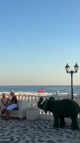 Two women are sitting on a white bench on a tiled patio. One woman is wearing a white top and white pants, and the other is wearing a black top and white pants. They are facing each other, and the woman in black is holding a phone. There is a green elephant statue to the right of the bench. In the background, there is a white railing with columns, the ocean, and a red boat on the water. The sky is clear and blue.