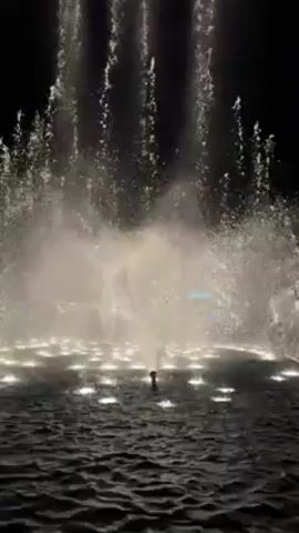 A water fountain display at night consists of a pool of water with multiple small lights illuminating the water jets. The water jets shoot up in various directions and heights, creating a dynamic spectacle. The background is dark, emphasizing the illuminated water. There's a single audible shout nearby.