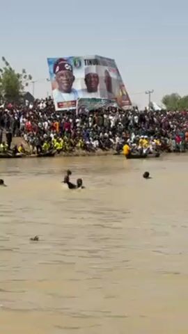 The video shows a large crowd gathered on the bank of a river, with people swimming in the water. There are several small boats in the water near the shoreline. In the background, there is a large billboard featuring the faces of two men, labeled with the name "TINUBU". The scene is captured in daylight under a clear sky. The water appears murky.