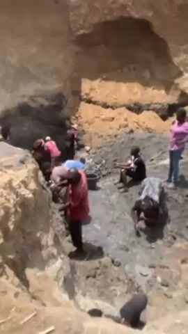 The video shows a group of people working in an excavated area, possibly a mine or quarry. Most are digging, shoveling, or moving material, possibly mud or clay. The excavated area has walls of yellow and brown earth, with the excavated material being dark gray. People are standing around, some are digging, some are sitting, and others are moving. The sky is visible and the weather appears to be clear.
