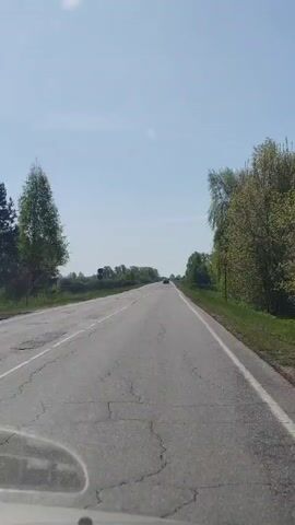 The video shows a view from inside a car, looking out through the windshield at a straight, two-lane road. The road has some cracks and a white line marking the right edge. The sky is blue with a few clouds. There are trees and green vegetation on either side of the road. In the distance, another car is visible on the road ahead.