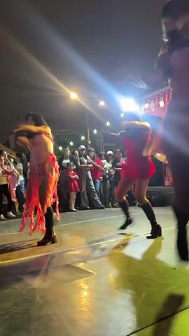A group of dancers perform on a stage at night. The dancers are wearing colorful outfits and the stage is lit with bright lights. There is an audience watching the performance.