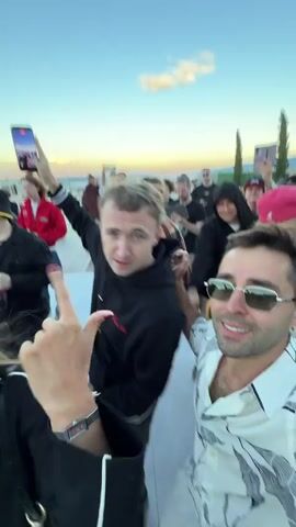 This media file captures a group of people outdoors during what appears to be a gathering or celebration. The subjects are a diverse group of individuals, with a focus on a few men in the foreground. One man, wearing sunglasses and a patterned shirt, is looking directly at the camera and appears to be speaking or reacting enthusiastically. Another man to his left, wearing a black hoodie, is smiling and holding up a phone, likely taking a selfie or recording a video. Other people are visible in the background, some with their faces blurred due to motion.

The setting is outdoors, with a clear sky suggesting it is daytime, likely late afternoon or early evening given the soft lighting. In the distance, there are hints of buildings and possibly a landscape, consistent with a location like Lisi, Georgia, which is known for its lake and recreational areas. The overall atmosphere seems lively and social, with people interacting and enjoying themselves. The presence of phones suggests the event is being documented.

There is no visible text in the media file. The emotions conveyed are primarily positive, with smiles and engaged expressions. The weather appears to be pleasant, suitable for an outdoor event.