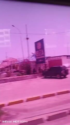 This is a video taken from inside a vehicle, likely a bus or car, as evidenced by reflections on the glass. The scene is dominated by a bright pink or purple hue, suggesting a filter or unusual lighting conditions.

The subjects include a few people walking on a sidewalk in the distance. There are also several objects visible: buildings with thatched roofs, lampposts, a prominent sign that reads "IBIZA" in large, dark letters, and yellow curbs along the roadside. The setting appears to be a semi-urban or coastal area, with what looks like a construction zone or undeveloped land to the right. The sky is a vibrant purple.

The activity taking place is simply passing by a street scene. The time of day is unclear due to the filtered colors, but the bright sky suggests it's daytime. The weather is likely clear, as there are no visible clouds or rain. There are no strong emotional cues conveyed due to the nature of the footage and the filtering. The text "Infinix HOT 50Pro" is visible in the bottom left corner, indicating the device used to record the video. The location is identified as Caraballeda, Venezuela.