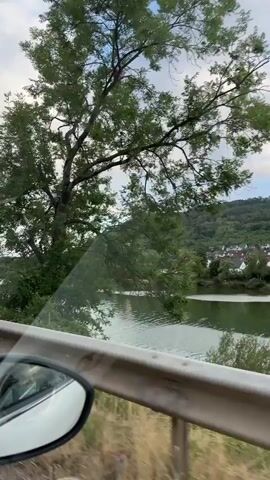 The video shows a view from inside a car, looking out the side window as the car drives along a road next to a body of water. The water appears to be a river or lake, with lush green trees and a hillside covered in vegetation on the opposite bank. A small town with houses and buildings is visible nestled on the hillside. The sky is partly cloudy with blue patches. A metal guardrail runs along the side of the road in the foreground, and the side mirror of the car is visible on the left. The camera is moving, creating a sense of motion as the scenery passes by.