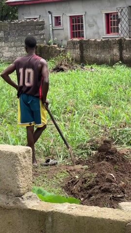 A person wearing a maroon tank top with the number 10 on the back, blue and yellow shorts, and flip-flops is standing in a grassy yard next to a pile of dirt. They are holding a long-handled tool, possibly a hoe or shovel. The person is facing away from the camera, with their left hand on their hip. Behind them, there is a house with a grey exterior and red window frames. The yard is filled with tall grass and some scattered debris. The foreground includes a concrete block wall. The camera angle is slightly low, looking up towards the person and the house. The focus of the video is variable, with the foreground initially sharp and then blurring as the camera adjusts to focus on the subject.