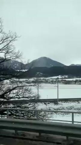 The view from a vehicle shows a winter landscape with snow-covered fields, trees, and mountains in the background under a gray sky. There is a highway with a metal guardrail to the lower left. Railroad tracks are visible closer to the snow covered landscape. There is a sign with a red arrow on a yellow background. Buildings are visible as the vehicle passes through a populated area.