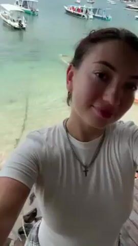 A woman with dark hair pulled back, wearing a white t-shirt and a silver chain necklace with a cross pendant, is taking a selfie video. She is standing on a wooden deck or platform overlooking a body of water. The water is a light turquoise color. In the background, there are several small boats anchored in the water. The sky is overcast. She initially looks directly into the camera, then lifts her hand and runs it through her hair. She continues to look at the camera with a slight smile. The camera angle changes slightly throughout the video as she holds the camera herself.