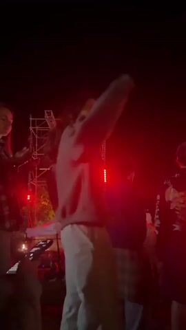 A nighttime outdoor scene shows a crowd of people in front of a stage structure with red lights. In the foreground, a person in a light-colored sweatshirt and pants is dancing with their arm raised. To their right, another person in a dark hoodie with a gold and black design is visible. Behind them, more people are gathered. The overall atmosphere appears to be that of a concert or outdoor event.