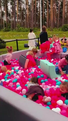 A vertical-format video shows a large, rectangular ball pit filled with hundreds of pink, white, and aqua-colored plastic balls. Several children play inside the pit, crawling, sitting, and throwing balls. Some soft, light-blue climbing structures are also in the pit. The ball pit is outdoors, on a grassy area surrounded by tall trees. In the background, adults sit on beanbag chairs, watching the children. A few vehicles are visible on a paved road in the distance. The weather appears overcast.