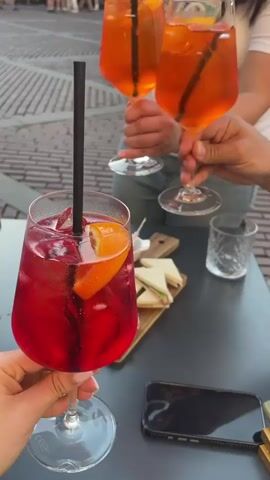 Here's a description of the video:

The video shows a group of people clinking glasses in a toast at an outdoor table. The glasses contain colorful cocktails with ice, straws, and orange slices. Some of the drinks are red, and others are orange. The table also holds a wooden cutting board with finger sandwiches, small bowls of olives, peanuts, and meatballs. A mobile phone and a lighter are also on the table.

The hands holding the glasses belong to multiple individuals, some with red nail polish. In the background, other people are walking on a paved area, suggesting an outdoor cafe or restaurant setting. Another person takes a photo of the drinks with a phone.