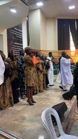A group of people are standing in a room, possibly a church or meeting hall, in front of a banner with text that reads "VISION STATEMENT" and "MISSION STATEMENT." The people are arranged in a line, with some holding microphones and singing. The individuals are dressed in colorful, traditional African clothing, and the women are wearing headscarves that match their outfits. The room has a light-colored floor and white pillars, with curtains in the background. There are also audio speakers visible in the foreground. The overall atmosphere appears to be a performance or a religious service.