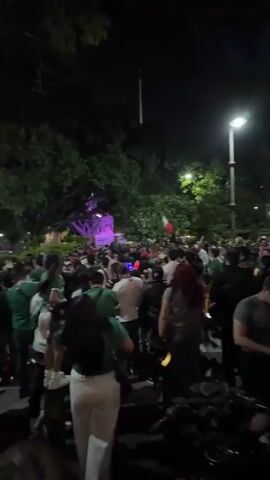 The video shows a crowded outdoor scene at night, possibly a public square or street, with many people gathered. Most individuals are wearing green or white, and several Mexican flags are visible, suggesting a celebration or event related to Mexico. The lighting is dim, with streetlights illuminating parts of the area. The crowd appears to be moving and interacting, with some individuals walking and others standing in groups. Motorcycles are parked in the foreground and among the crowd. The overall atmosphere seems festive.