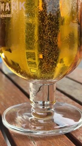 The video shows a close-up shot of a glass of beer. The beer is golden in color and has many small bubbles rising to the surface. The glass has some condensation on the outside. The brand name "Tuborg" and "MARK" are visible on the glass. The beer is sitting on a wooden table. The background is blurred, but it appears to be an outdoor setting with some reflections in the glass, possibly of buildings or lights.