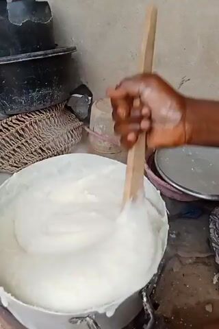 A person is seen stirring a large pot of white, pasty food with a wooden spoon. The pot is aluminum, and the person's hand is dark-skinned. A wicker basket, a black container, and a pink dish are visible in the background. The environment appears to be a simple kitchen.