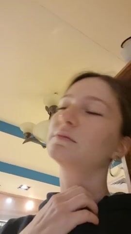 A woman in a black top and hoop earrings looks at the camera and places her hand on her neck and cheek. The scene is indoors. In the background is a shelf with objects on it and a light fixture. The ceiling is a pale yellow color.