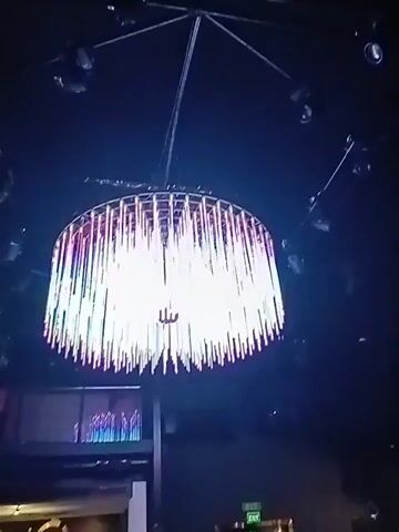 A low-angle shot shows a large, modern chandelier hanging from the ceiling. The chandelier is round and tiered, with many strands of light that hang down like fringe. The strands change color, shifting from white to green to blue to purple. Around the chandelier are many spotlights aimed down. The background is dark, with a glimpse of a window and some furniture visible below the chandelier.