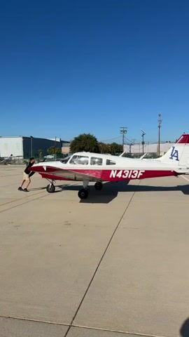 A young person is pushing a small white and red airplane. The airplane has the registration number N4313F printed in white on its red tail. The person is wearing a dark t-shirt and shorts. They are pushing on the nose of the airplane. The airplane is on a concrete surface, likely an airport tarmac, with power lines and buildings in the background. The sky is clear and blue.