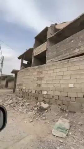 A vehicle drives along a dirt road past buildings. The buildings are mostly made of concrete block and appear weathered or partially destroyed.  Power lines run overhead. The video is taken from inside the vehicle.  There are no people visible. The environment appears dusty and dry.