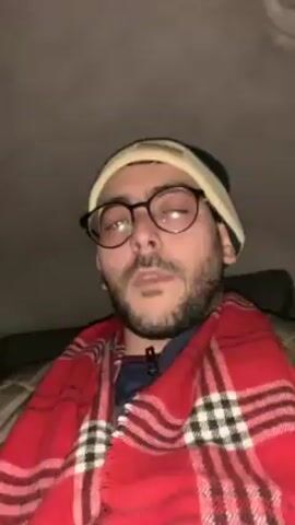 A man is shown in a car. He has a beard and wears glasses, a beanie, and a red and white plaid scarf. The car interior is dark. The man looks up with a neutral expression as music plays.