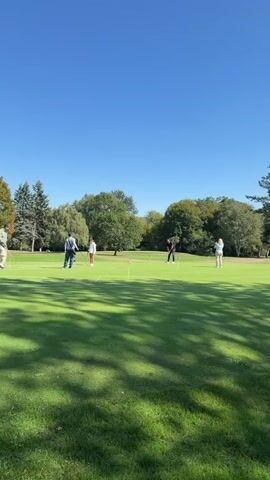 The video shows a group of people playing croquet on a sunny day on a well-maintained lawn with trees in the background. The camera pans across the scene, showing several people hitting croquet balls with mallets, aiming for wickets. One person is visible in the foreground on the left, another in the middle distance on the left, and two people are in the middle distance on the right. In the background, several other people can be seen walking along a path beside the croquet lawn. The sky is clear blue and the sun is shining, casting shadows across the grass.