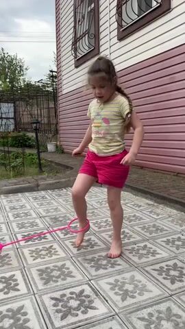 A young girl with light brown hair pulled back is outdoors playing a jump rope game. She's barefoot on a patio made of large square tiles with a floral pattern, wearing a striped t-shirt and pink shorts. The jump rope toy is also pink, attached to her ankle and spinning. Behind her is a pink-sided house with a white section and a window with a metal grate.  A garden area with plants and a metal arch is visible in the background. The sky is overcast.