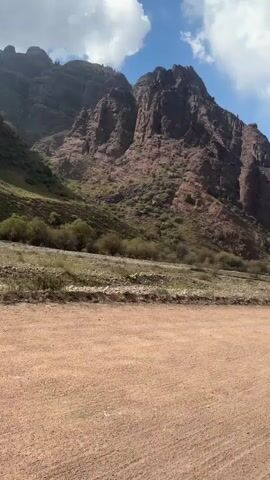 This is a first-person perspective video taken from a vehicle driving on a dirt road. The camera pans across a mountainous landscape with rocky, reddish-brown peaks and sparse green vegetation. The sky is blue with scattered white clouds. The road appears to be unpaved and dusty. The vehicle's white hood is visible at the bottom of the frame in some shots, indicating movement.
