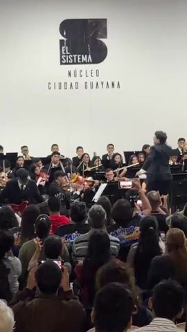 A wide shot of a live orchestra performing, viewed from the audience's perspective. The orchestra members, dressed in black suits, are playing a variety of instruments including violins, trumpets, and a guitar. A conductor is leading the orchestra. The audience is a mix of adults and children, many of whom are holding up their phones to record the performance.  Behind the orchestra, a large sign displays "EL SISTEMA NÚCLEO CIUDAD GUAYANA." The room is brightly lit. The atmosphere appears celebratory.