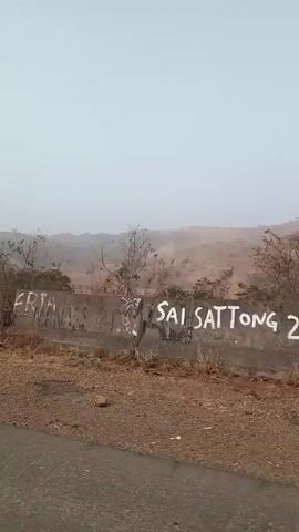 The video, seemingly taken from a moving vehicle, shows a landscape featuring a road, a low retaining wall, and a distant hill covered in sparse vegetation. The sky is overcast, suggesting a foggy or hazy atmosphere. Graffiti is visible on the retaining wall. The foreground consists of the asphalt road and a small shoulder area. In the midground, the retaining wall separates the road from a patch of dry vegetation. Beyond the wall, the hill rises in the background, its slopes covered in a mix of brownish grass and scattered trees. The overall impression is a rural or semi-arid landscape, potentially shot during a time of year when vegetation is less lush.